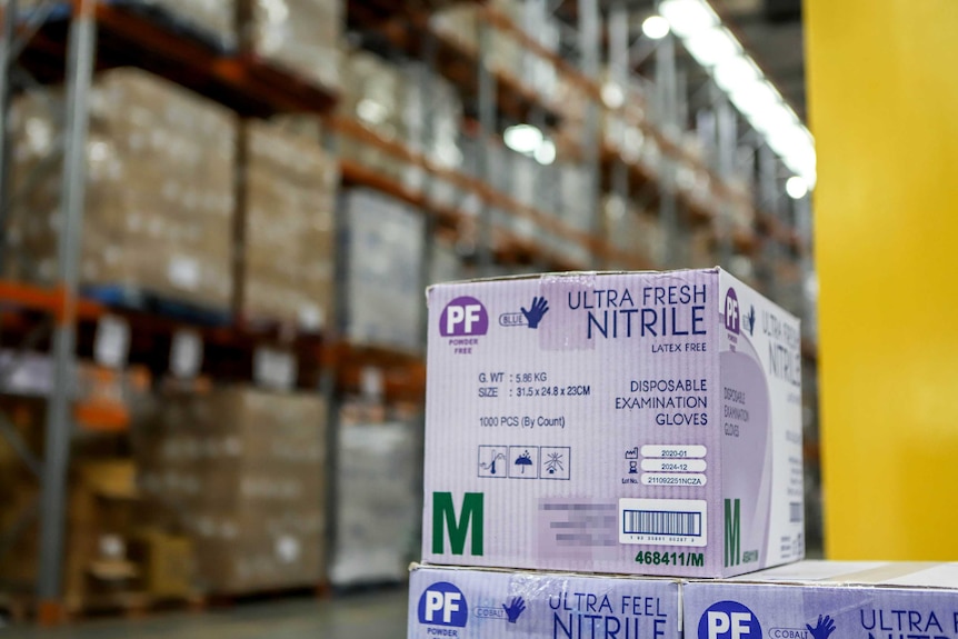 A purple box with disposable examination gloves inside sits in the foreground with large shelves of boxes blurred in backgrond.