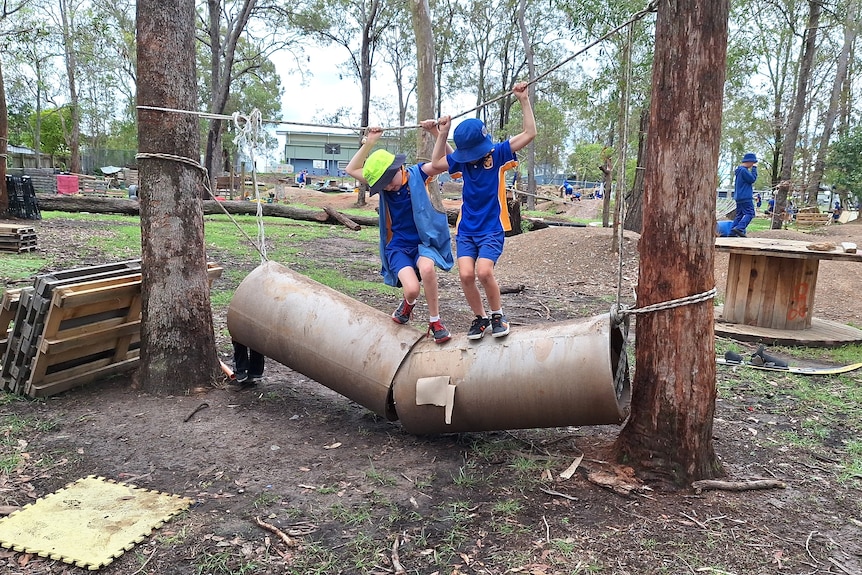 Two children grasp an overhead rope as they balance on slipper, mud-covered concrete tubes between two gum trees.