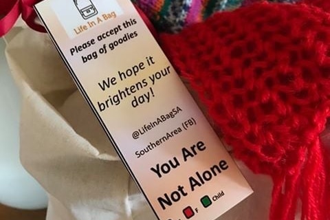 A label reading 'Please accept this bag of goodies, we hope it brightens your day. You are not alone!' and a knitted red scarf