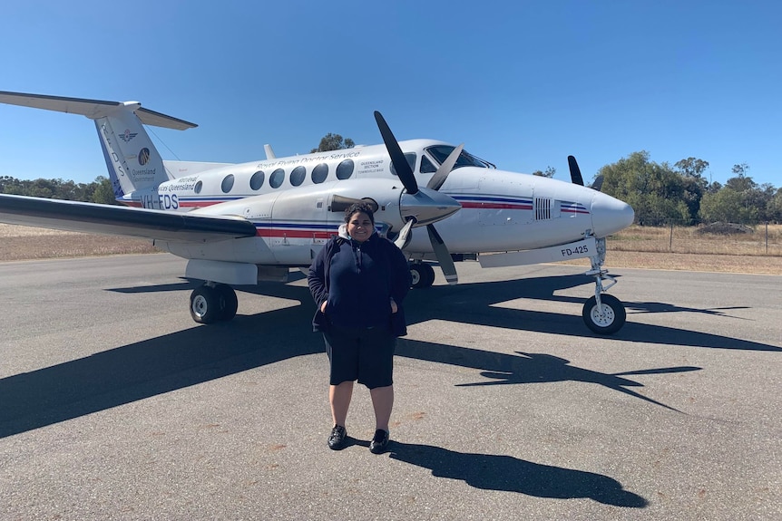 Chelsea standing on the tarmac with her hands in her pockets, royal flying doctors service plane behind her.