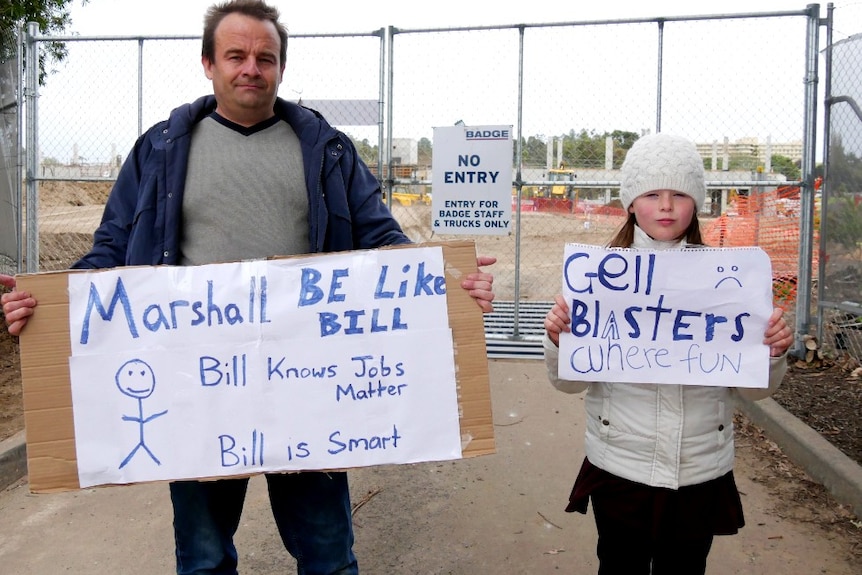 Man and young girl holding protest signs outside construction site.