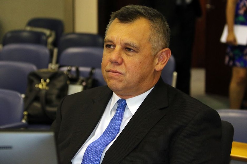 Indigenous man sits in a court room, with a laptop in front of him, and with a vibrant blue tie on.