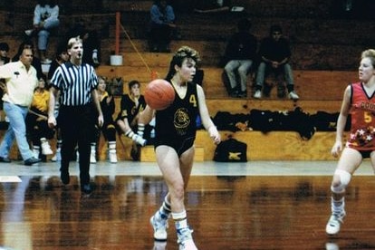 a teenage girl in basketball uniform dribbles the ball down the court. She has a wicked mullet with blonde tips over brown locks