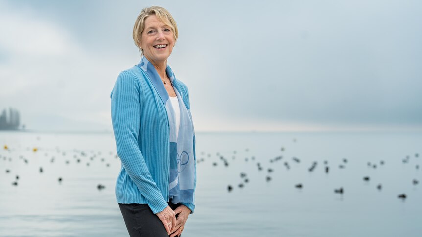 Michelle stands in front of water, side on, smiling. Birds sit on the water in the background.