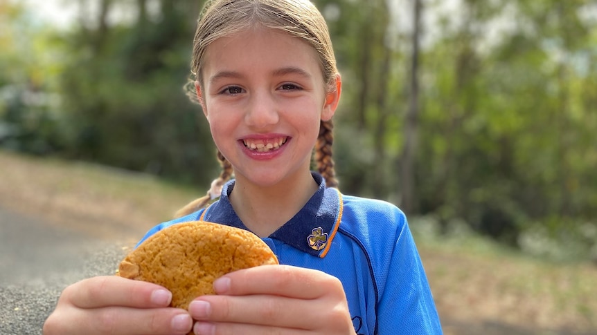 Young girl in Girl Guides uniform holds cookie and smiles with hair in two braids.
