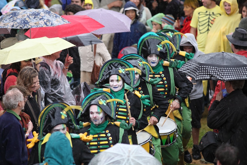 A troupe of drummers walks through a crowd of people with umbrellas.