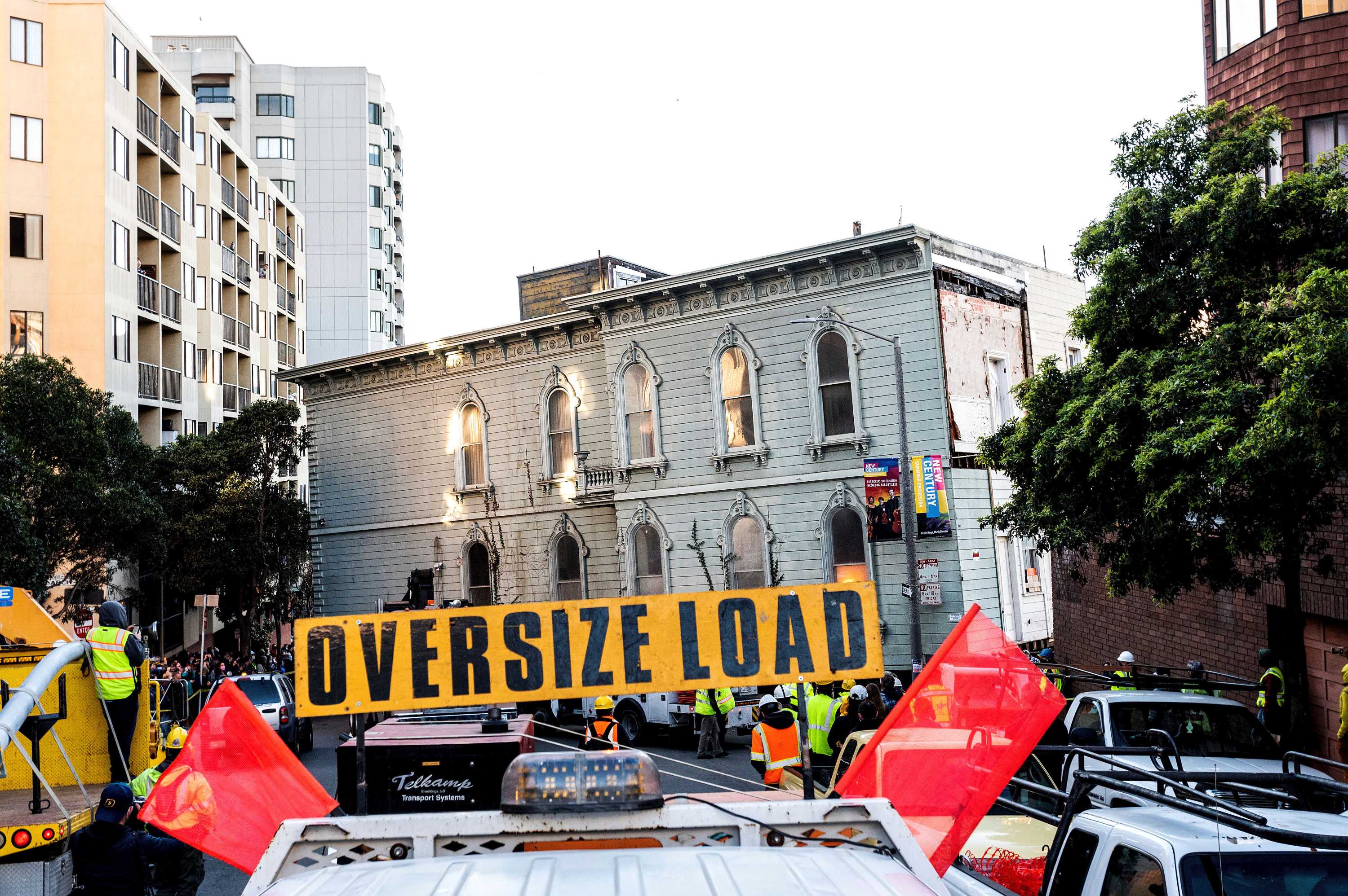 A light green house is seen in the middle of a street with tens of people watching. A sign warns: "Oversize load"