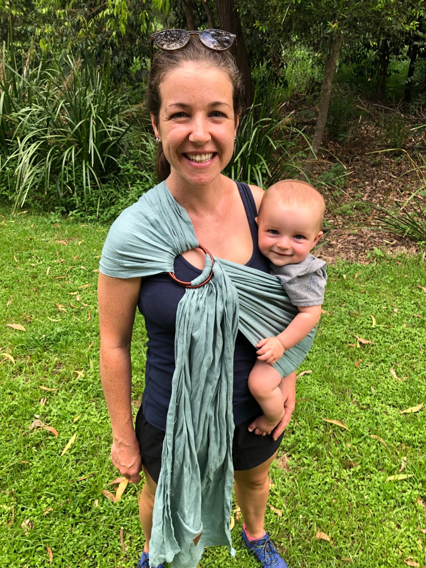 A woman smiles as she holds a baby, attached at her side in a big wrap. They stand in a grassed garden.