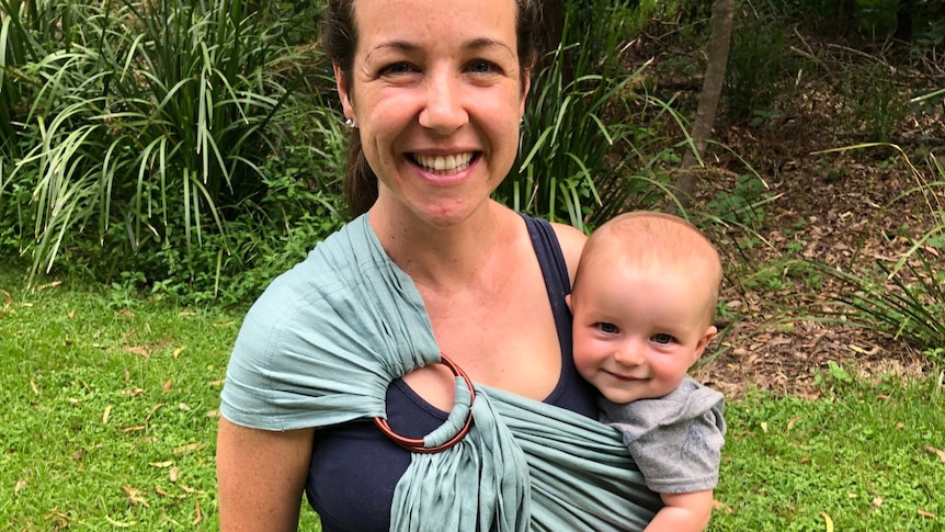 A woman smiles as she holds a baby, attached at her side in a big wrap. They stand in a grassed garden.
