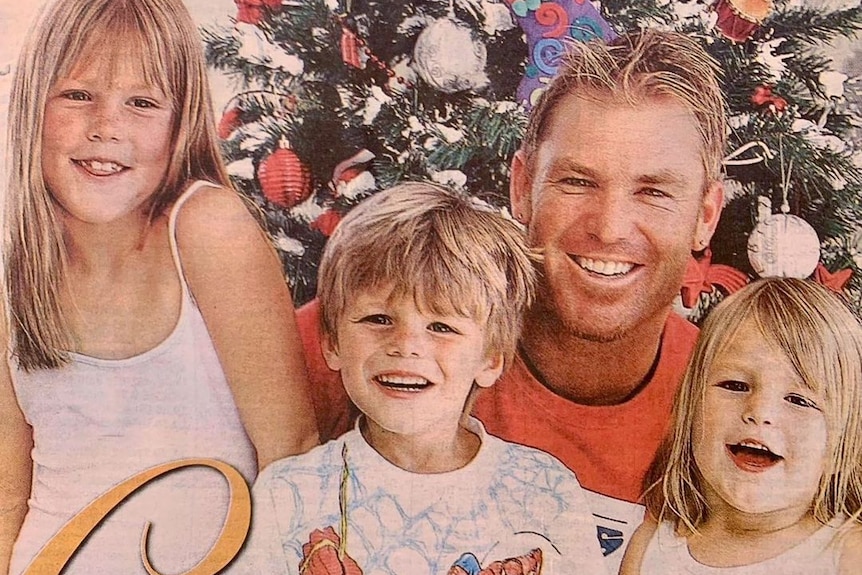 A retro colour photo of Shane Warne and his children smiling in front of a Christmas tree