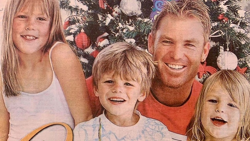 A retro colour photo of Shane Warne and his young children smiling in front of a Christmas tree