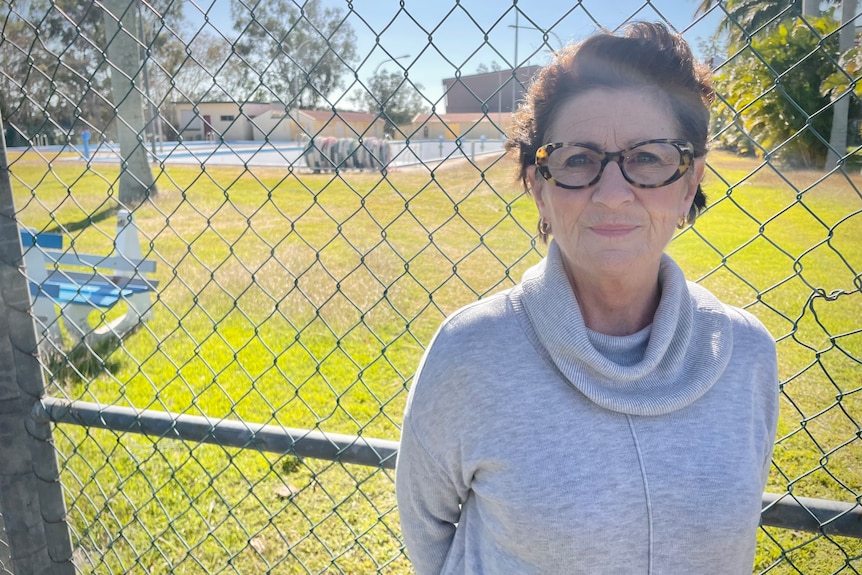 A woman with dark hair, wearing glasses, stands in front of a wire fence.