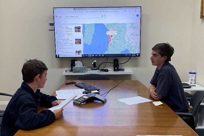 Two boys sit across a table from each other in a conference call with sponsors with a large monitor of Google maps in the back