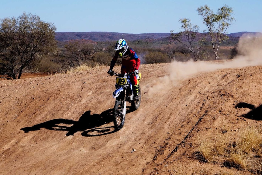 Photo of a boy riding a dirt bike down a red track with a dust trail.