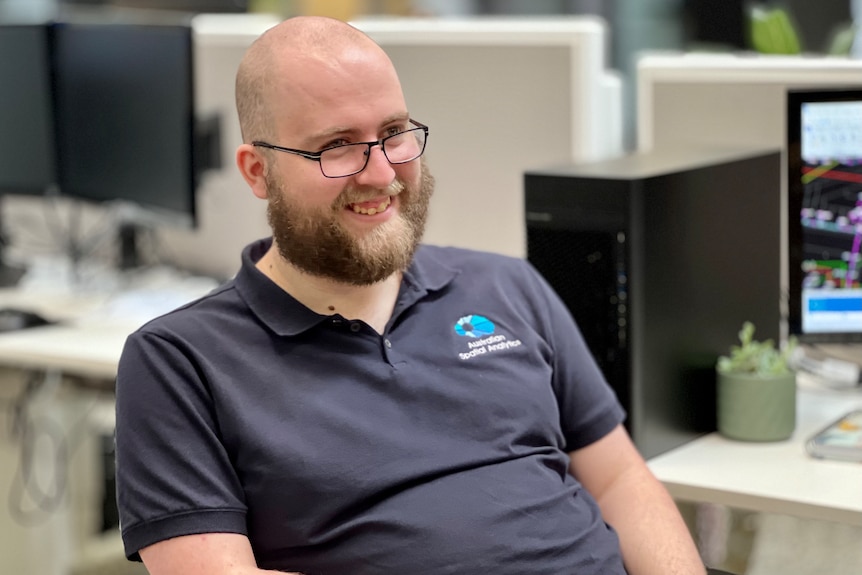 A bald man with a beard sits on an office chair and is smiling at someone off camera.