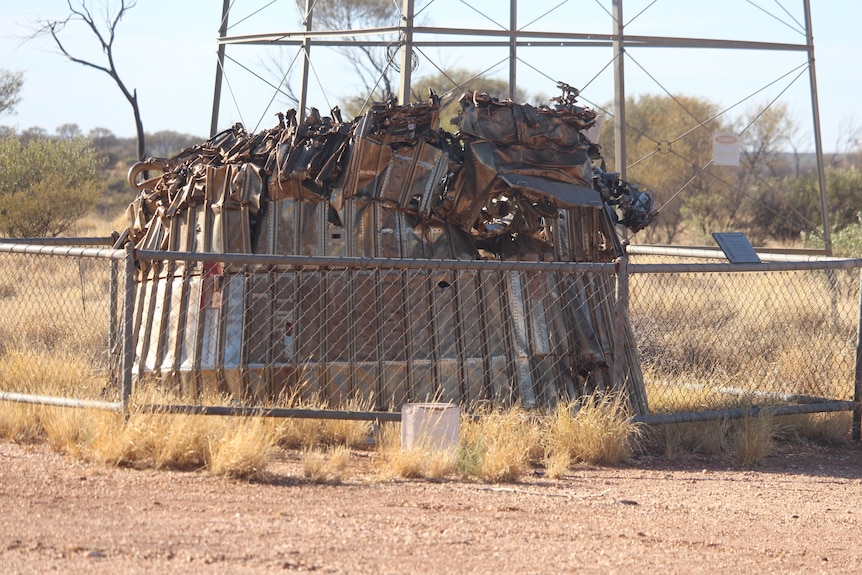 A crumpled mass of metal behind a fence, a metal tower behind, red earth, light blue sky.