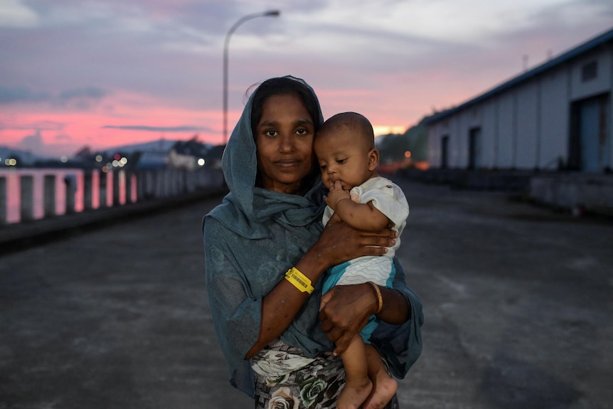 As the sun sets, a Rohingya woman stands in a compound, holding her baby. 