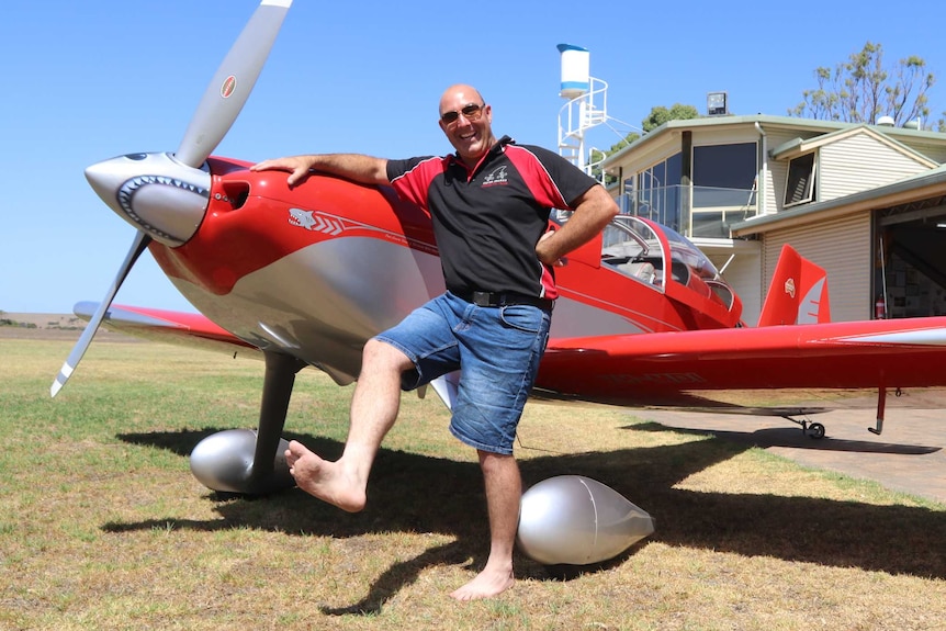 Red aeroplane on grass in front of hanger, Man holding foot up near front of plane