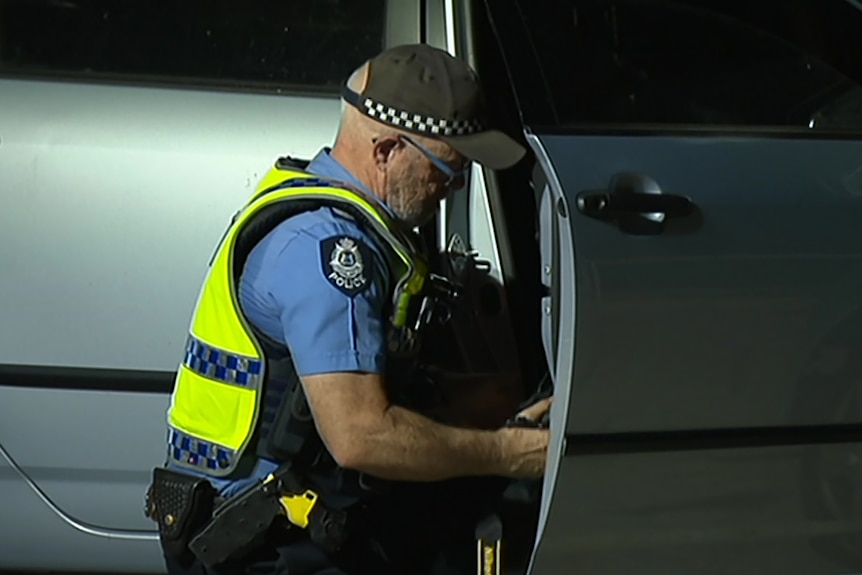 A close-up shot of a police officer examining the driver's side of a vehicle at night.