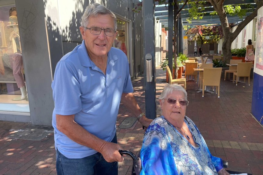 An older man with glasses and a polo shirt pushing an older woman in a wheelchair.