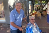 An older man with glasses and a polo shirt pushing an older woman in a wheelchair.