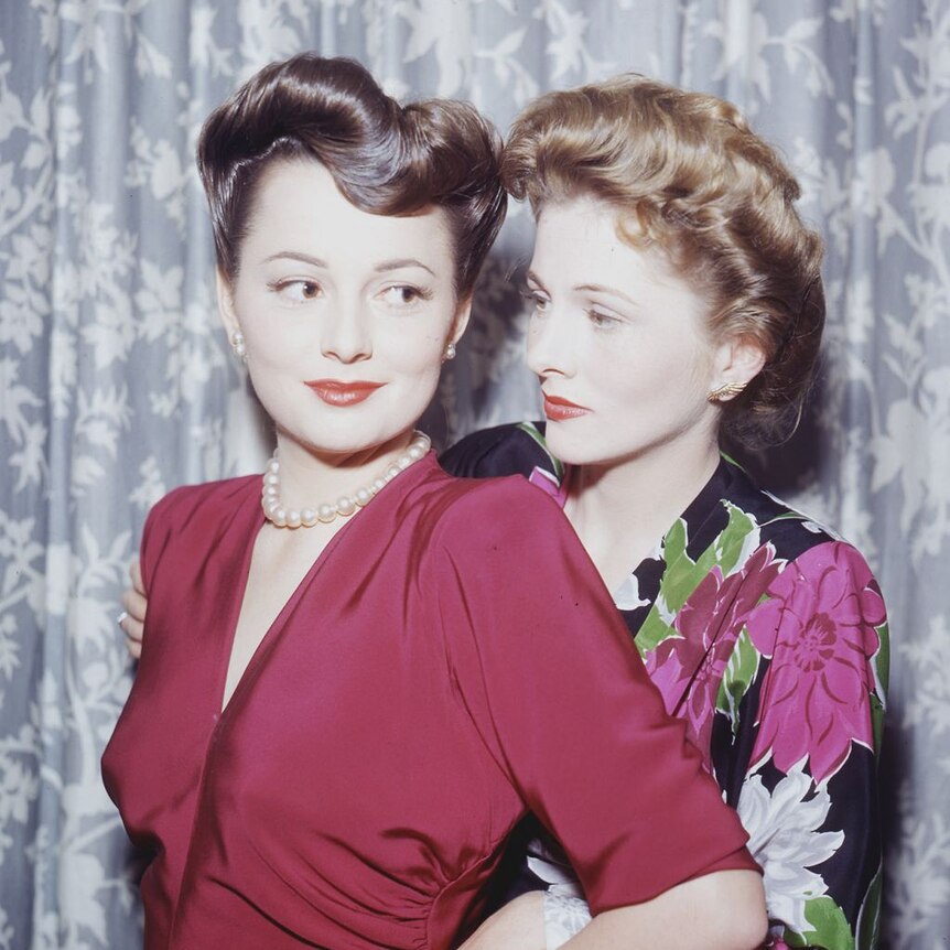 A vintage photograph of two sisters, one on the left wearing a dark-pink dress, the one on the right wearing floral.