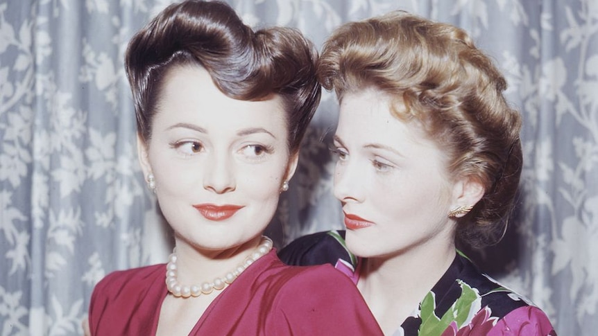 A vintage photograph of two sisters, one on the left wearing a dark-pink dress, the one on the right wearing floral.
