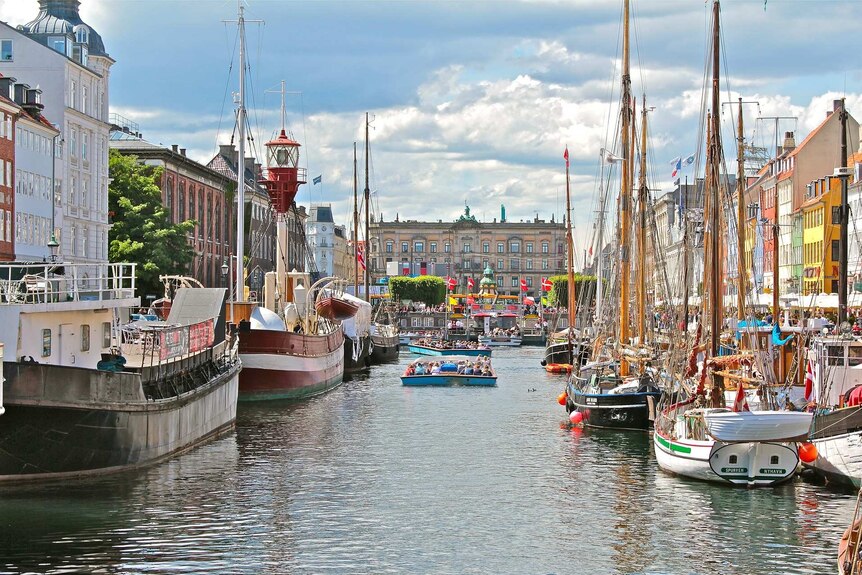 On a bright day, you view an old canal of Copenhagen, lined by heritage-era buildings and sail boats.