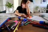 A boy with his arm covering his face sits at a desk with a pencil case spilling out pens and markers.