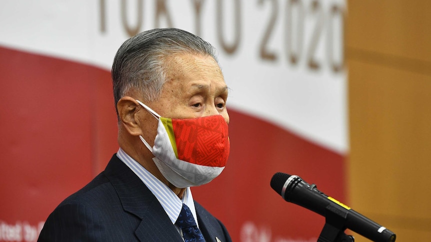 Man with grey hair stands in profile in front of a microphone wearing a red mask.
