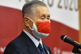 Man with grey hair stands in profile in front of a microphone wearing a red mask.