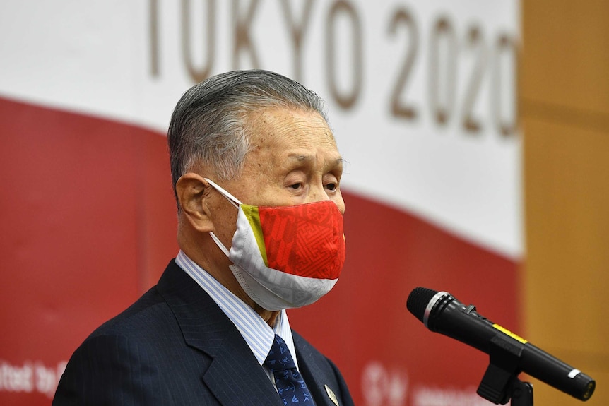 Man with grey hair stands in profile in front of a microphone wearing a red mask.