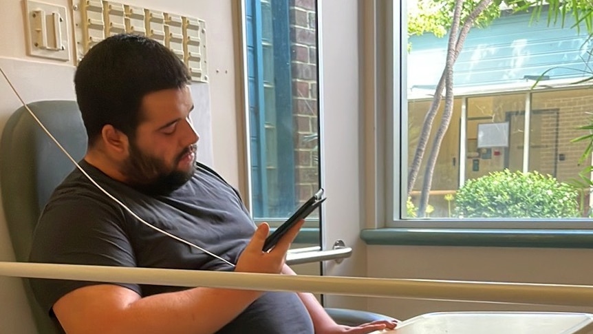 A young man is in a hospital chair and looks at his phone. There's a rail in front of him, he has a beard.