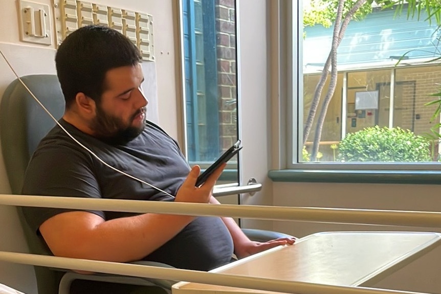 A young man is in a hospital chair and looks at his phone. There's a rail in front of him, he has a beard.