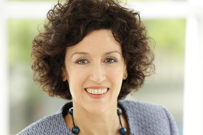 A professional headshot of a woman with brown curly hair and a suit jacket smiling at the camera