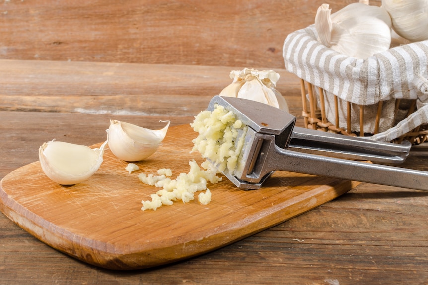 A chopping board with two garlic cloves, and a garlic crusher