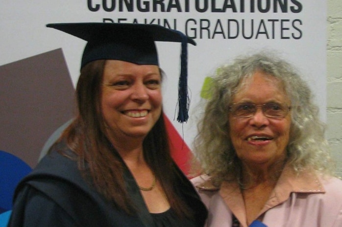 A young woman wears a graduation cap and stands smiling next to her mother .