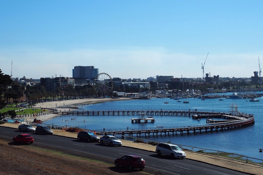 A circular boardwalk, bay beach, marina, ferris wheel and Geelong's city centre on a sunny day.