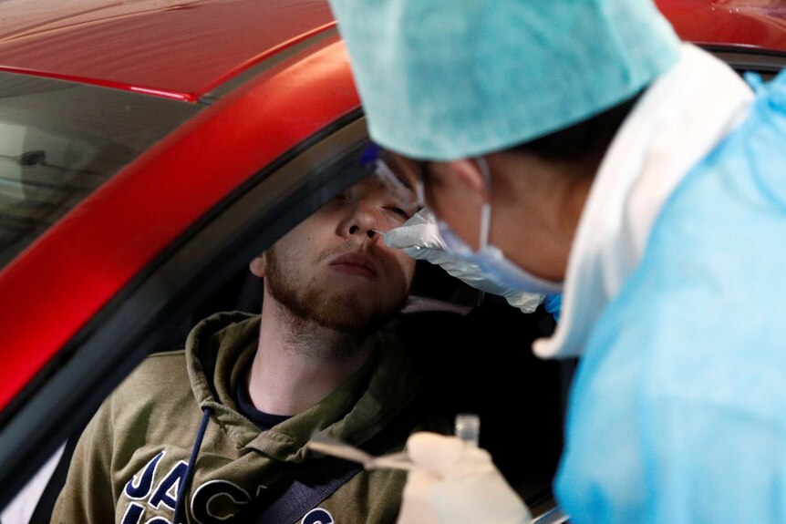 A man sits in a red car while a doctor in blue scrubs and face mask puts a small stick in his nose.