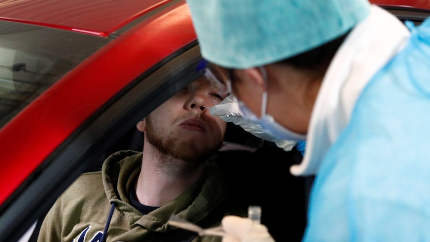 A man sits in a red car while a doctor in blue scrubs and face mask puts a small stick in his nose.
