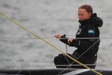 A wet-looking Greta Thunberg wearing black is sitting on a boat in the middle of the ocean, smiling she holds ropes of the boat