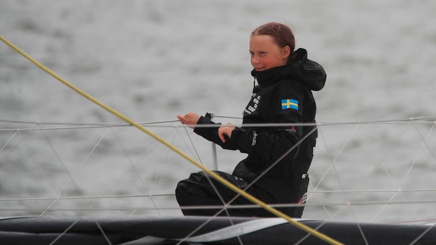 A wet-looking Greta Thunberg wearing black is sitting on a boat in the middle of the ocean, smiling she holds ropes of the boat