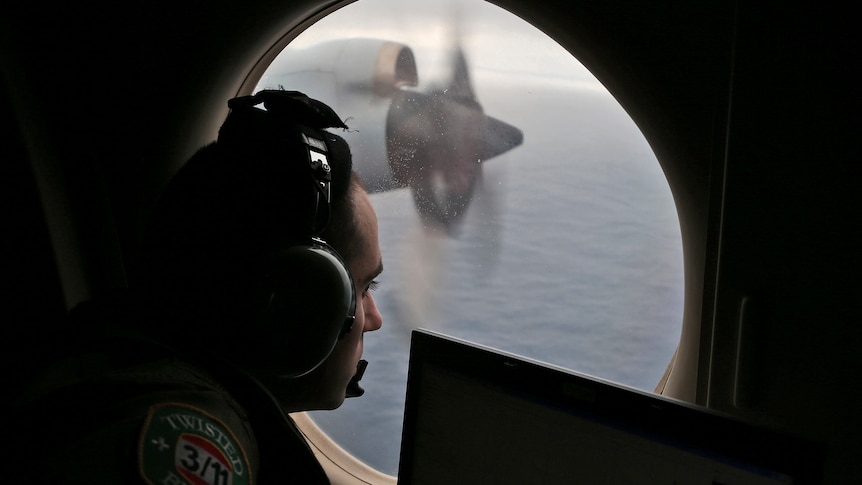 A man from an Air Force search team in a headset looks out a plane window looking at the ocean. 