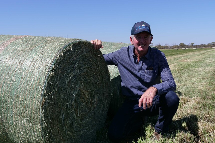 A man kneels next to a bale of hay and smiles at the camera