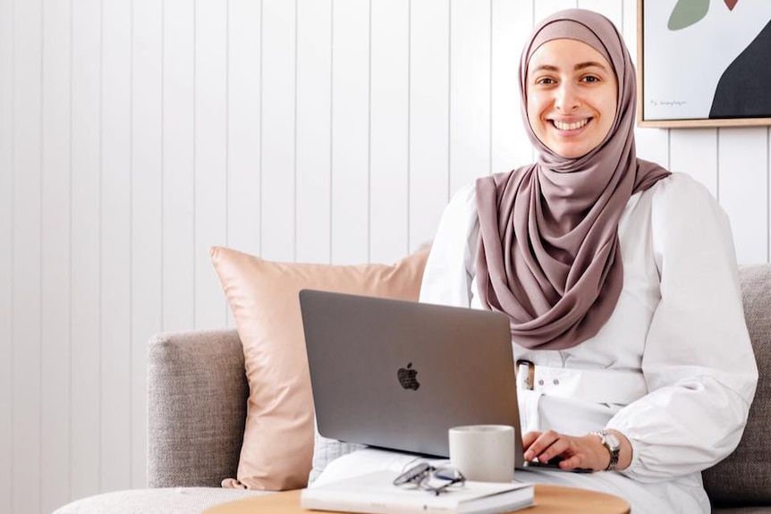 Mariam, who wears a headscarf, smiles on a couch, holding a laptop.