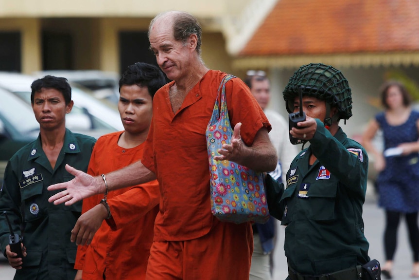 A man in an orange prison jumpsuit is escorted by Cambodian officers