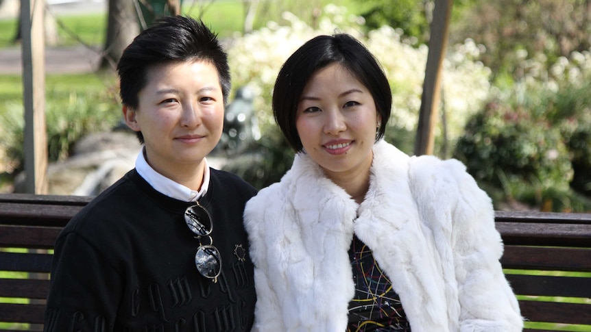 Tick Jiang and Joyce Zhang sit on a park bench and smile at the camera.