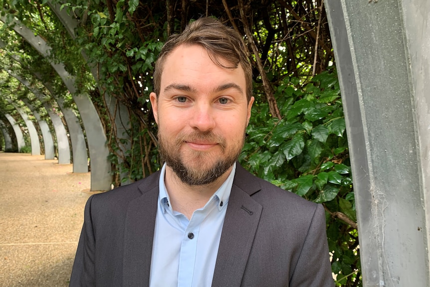 A man with brown hair and a beard wears a suit and smiles at the camera