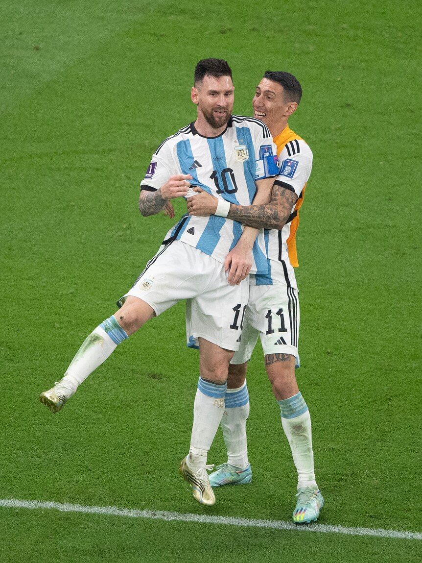 Two soccer players wearing white and blue hug on the grass during a game