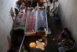 A group of people standing behind three coffins. A woman sits in front of the coffins holds a photo, mourning